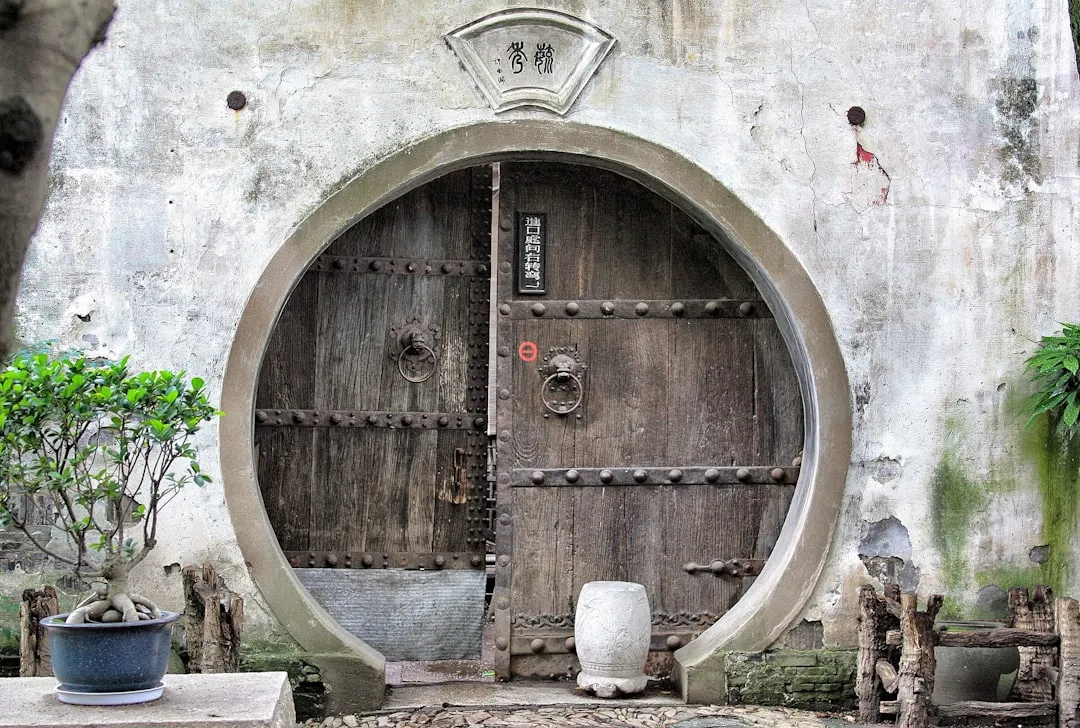 Portail en bois rond avec détails métalliques, dans une vieille muraille, décor zen avec plantes et poterie, ambiance asiatique, symbole de philosophie orientale, liés à la sagesse et la méditation.